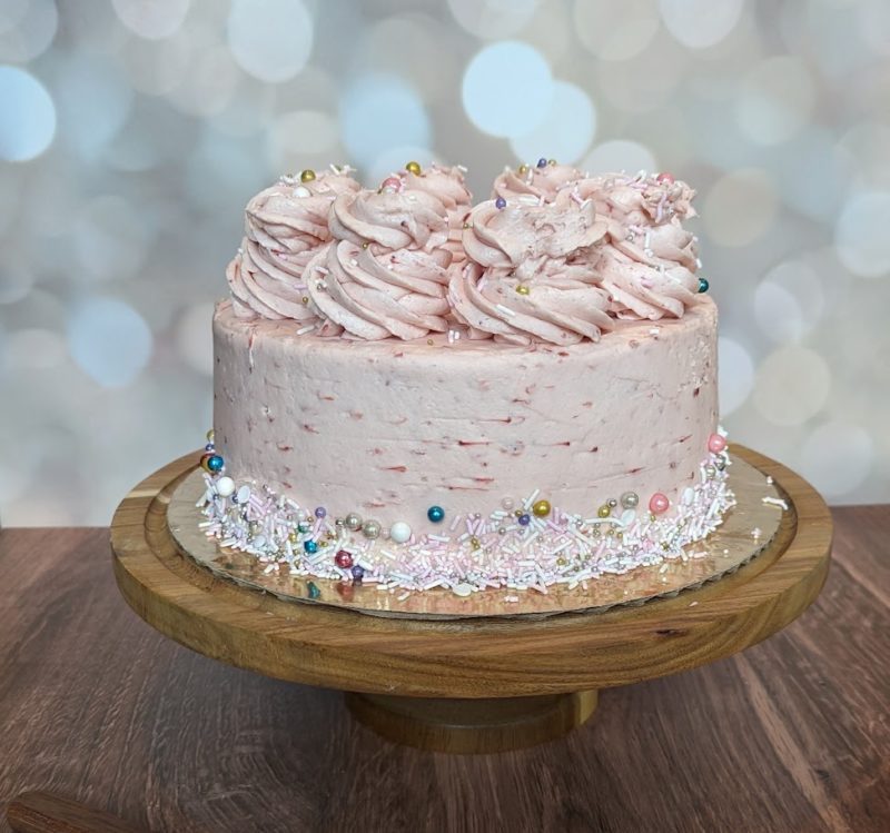 Strawberry pink layer cake with pink buttercream swirls on top and a band of multicolored sprinkles around the base and on top